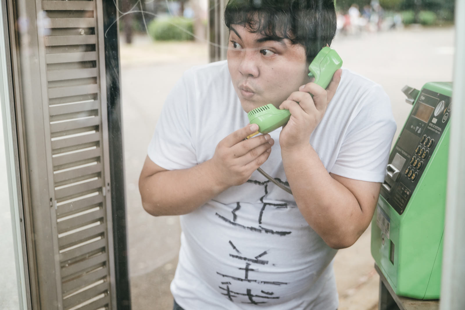緑色の公衆電話ボックスで受話器を持って通話している白いTシャツの男性［モデル：段田隼人］