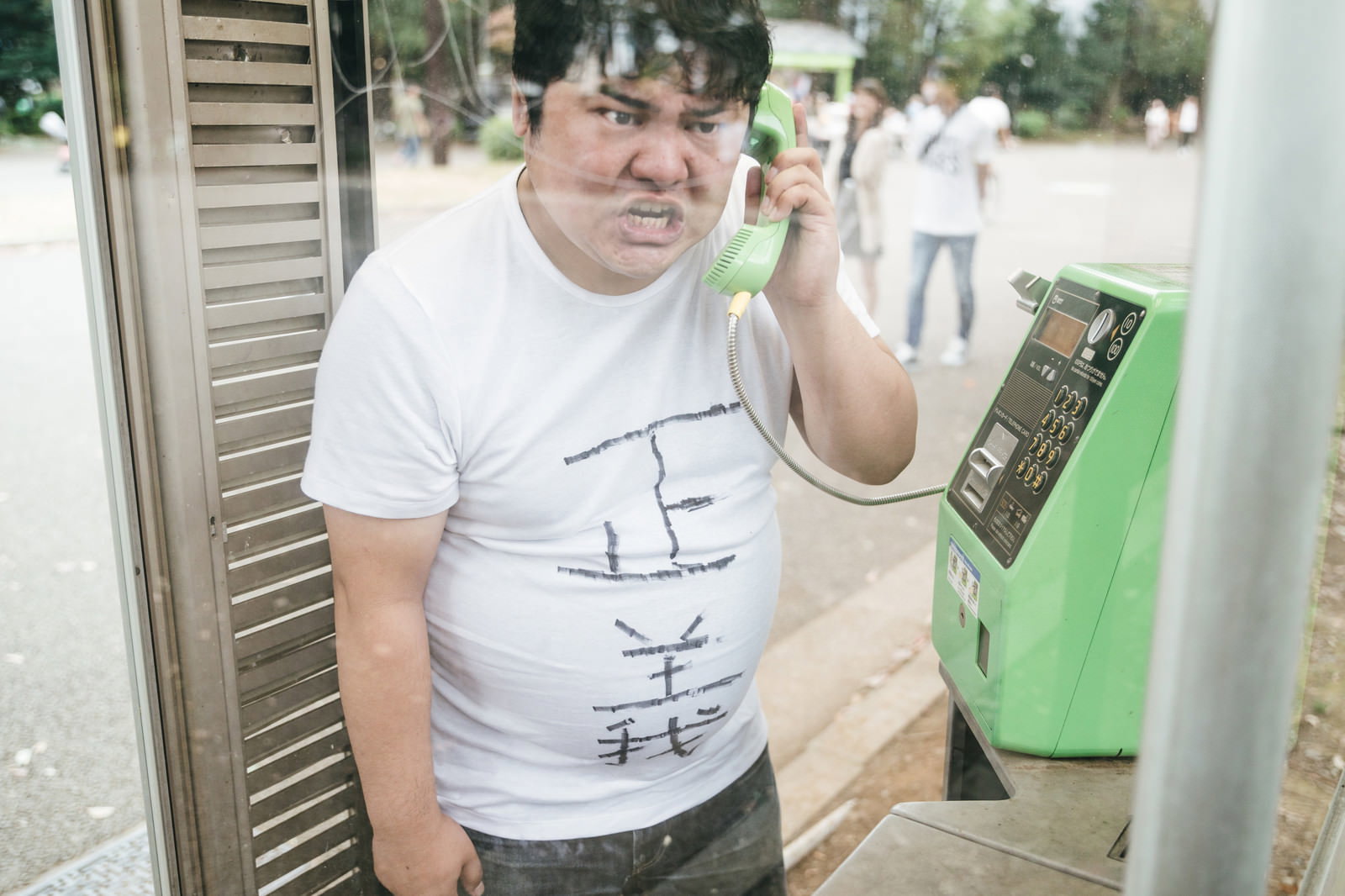 緑色の公衆電話で通話する白いTシャツの男性［モデル：段田隼人］