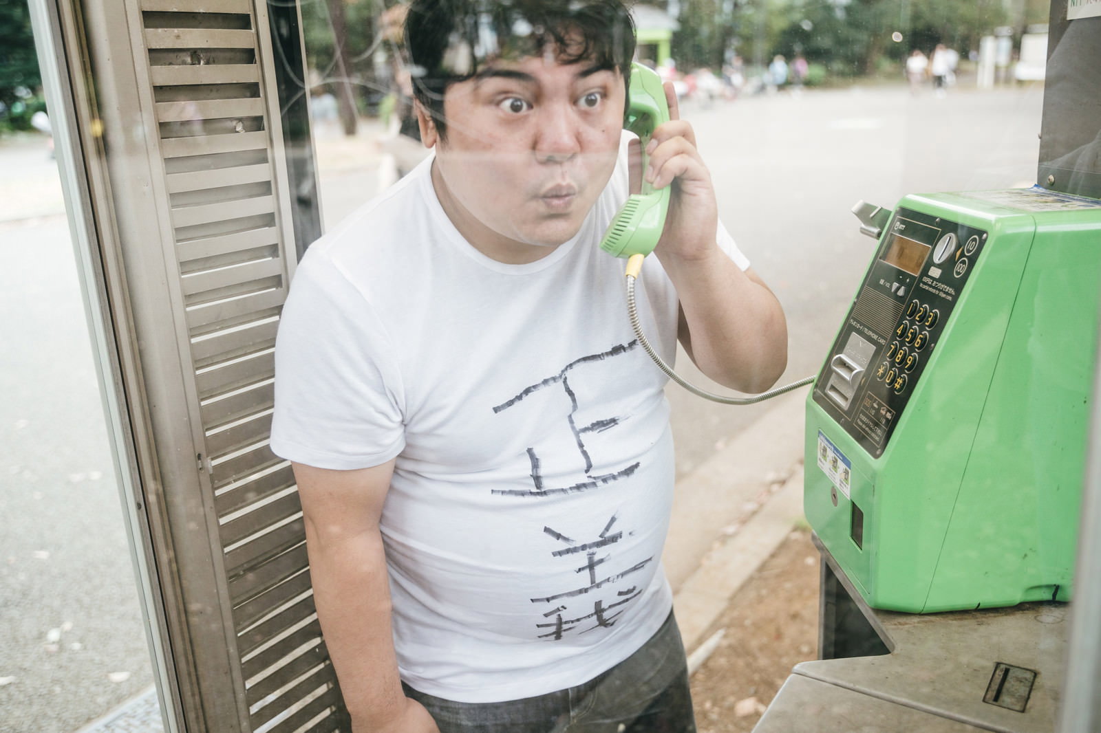 緑色の公衆電話で電話をかけている白いTシャツの男性［モデル：段田隼人］