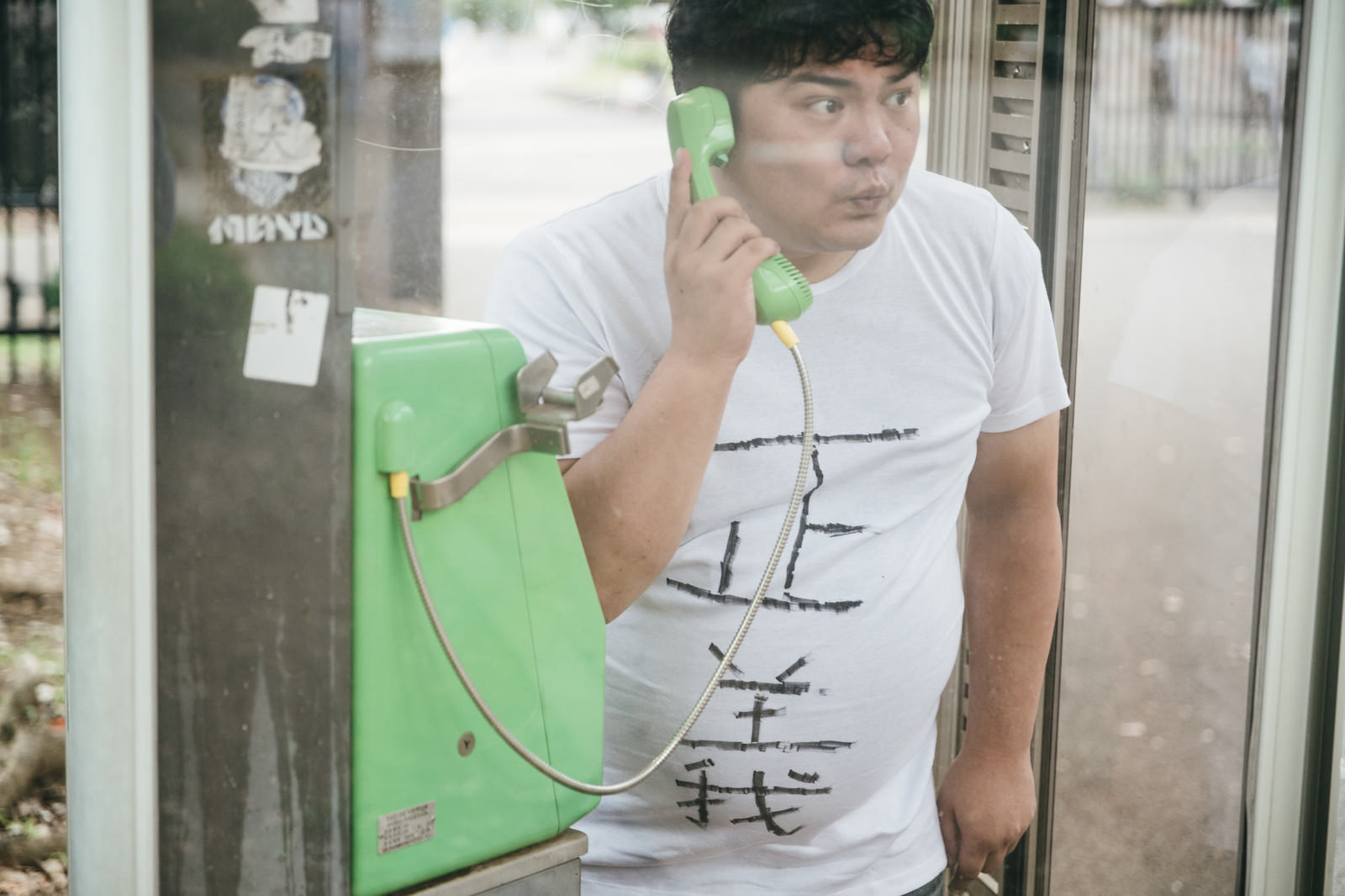 緑色の公衆電話で通話している白いTシャツの男性［モデル：段田隼人］