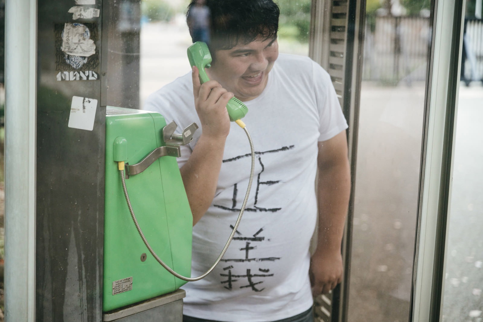 緑色の公衆電話で通話している白いTシャツの男性［モデル：段田隼人］