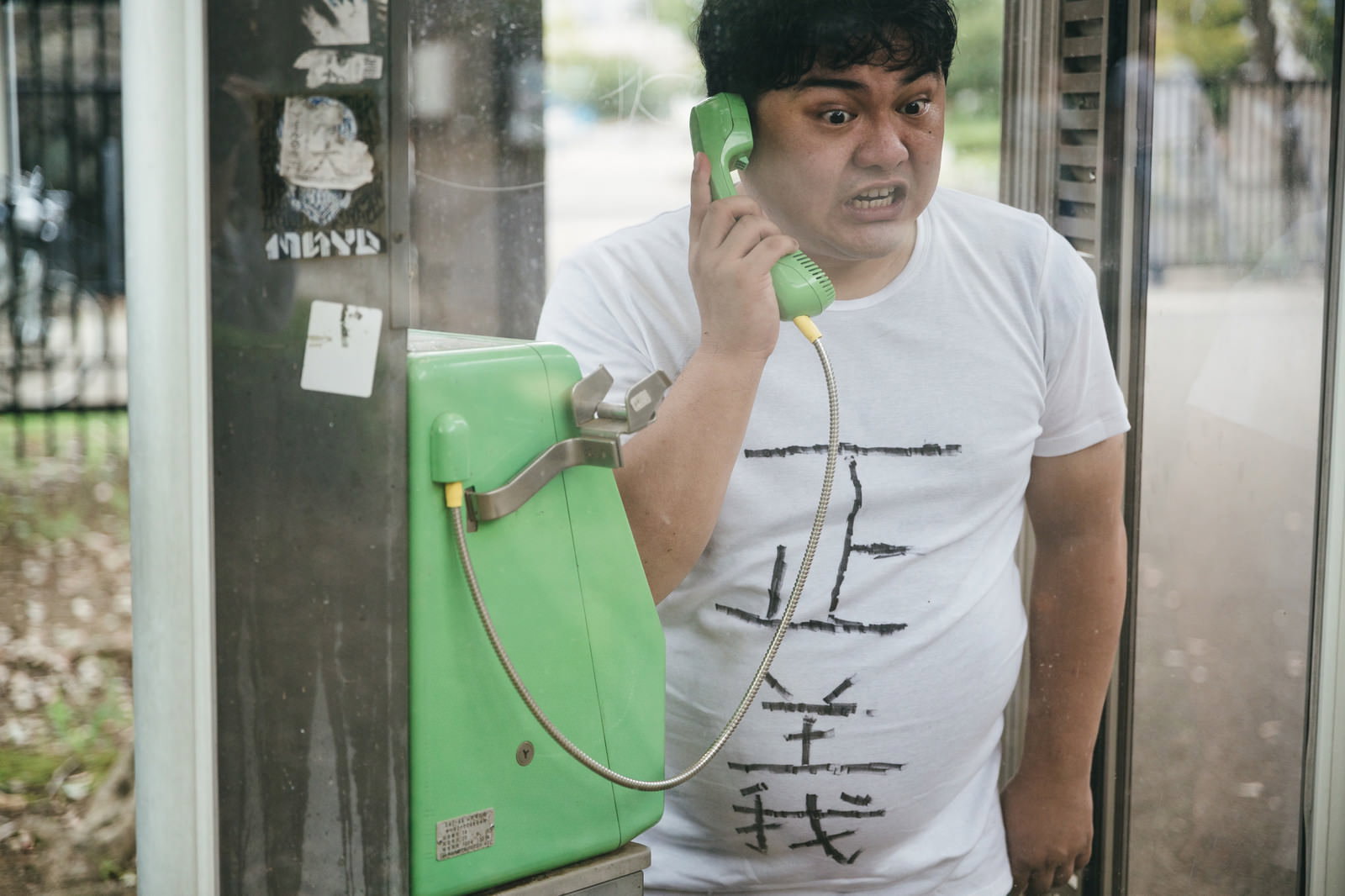 公衆電話の受話器を握り真剣な表情で通話する白いTシャツの男性［モデル：段田隼人］