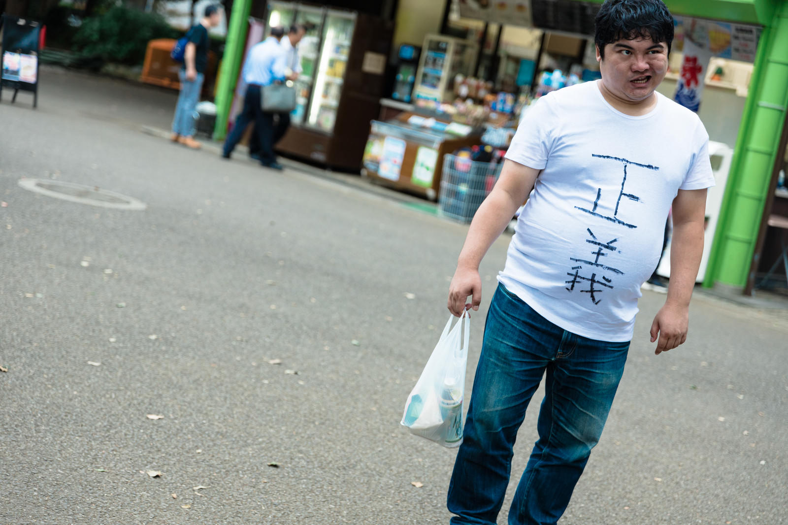 コンビニ前で不満そうな表情でテイクアウト袋を持つ白いTシャツの男性［モデル：段田隼人］