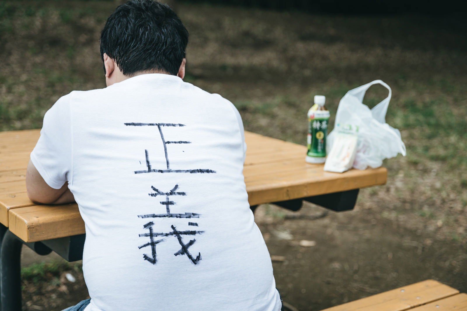 公園のベンチで弁当を食べる正義Tシャツの男性の後ろ姿［モデル：段田隼人］
