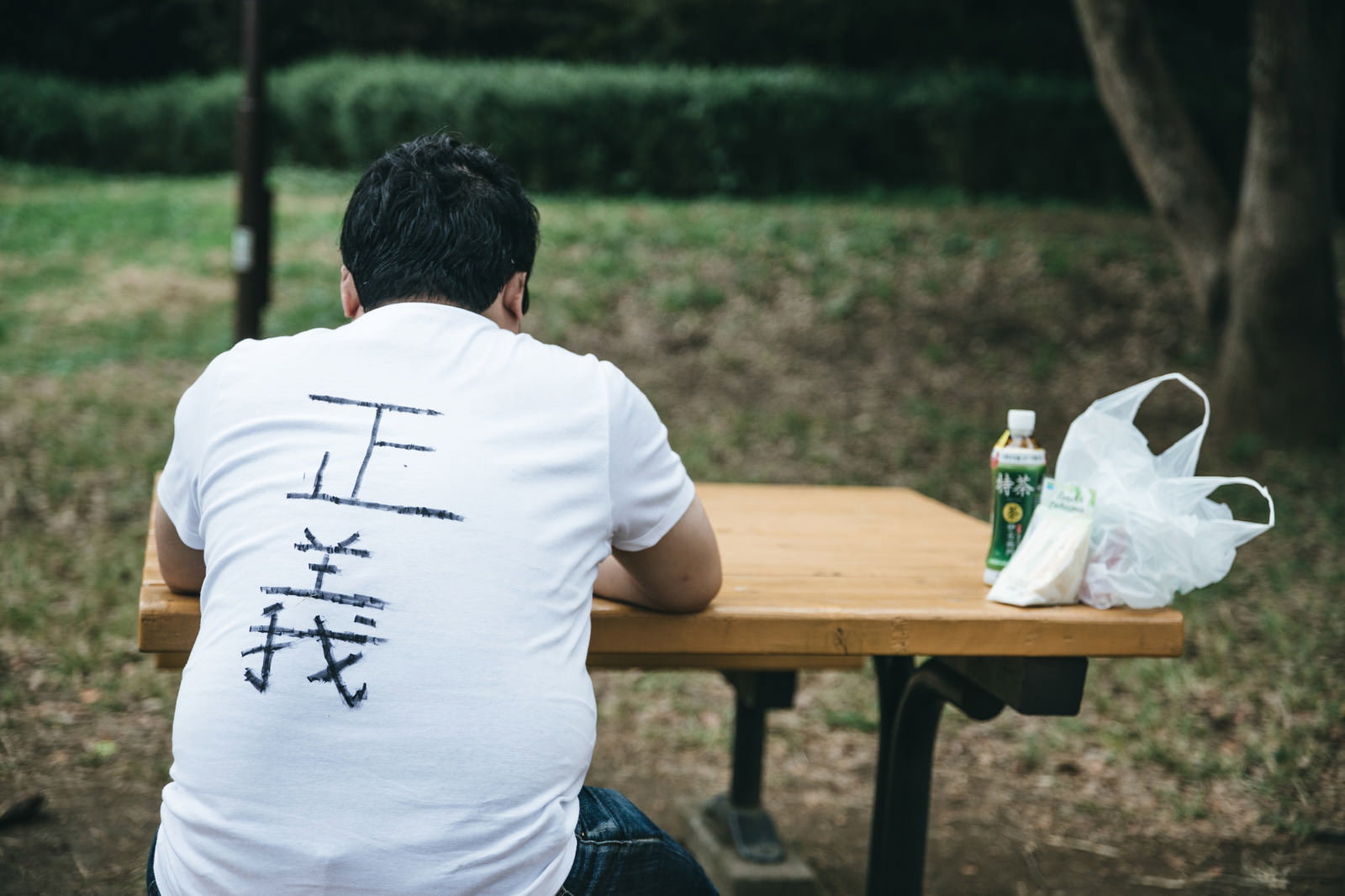 公園のベンチで一人食事をする白いTシャツの男性の後ろ姿［モデル：段田隼人］