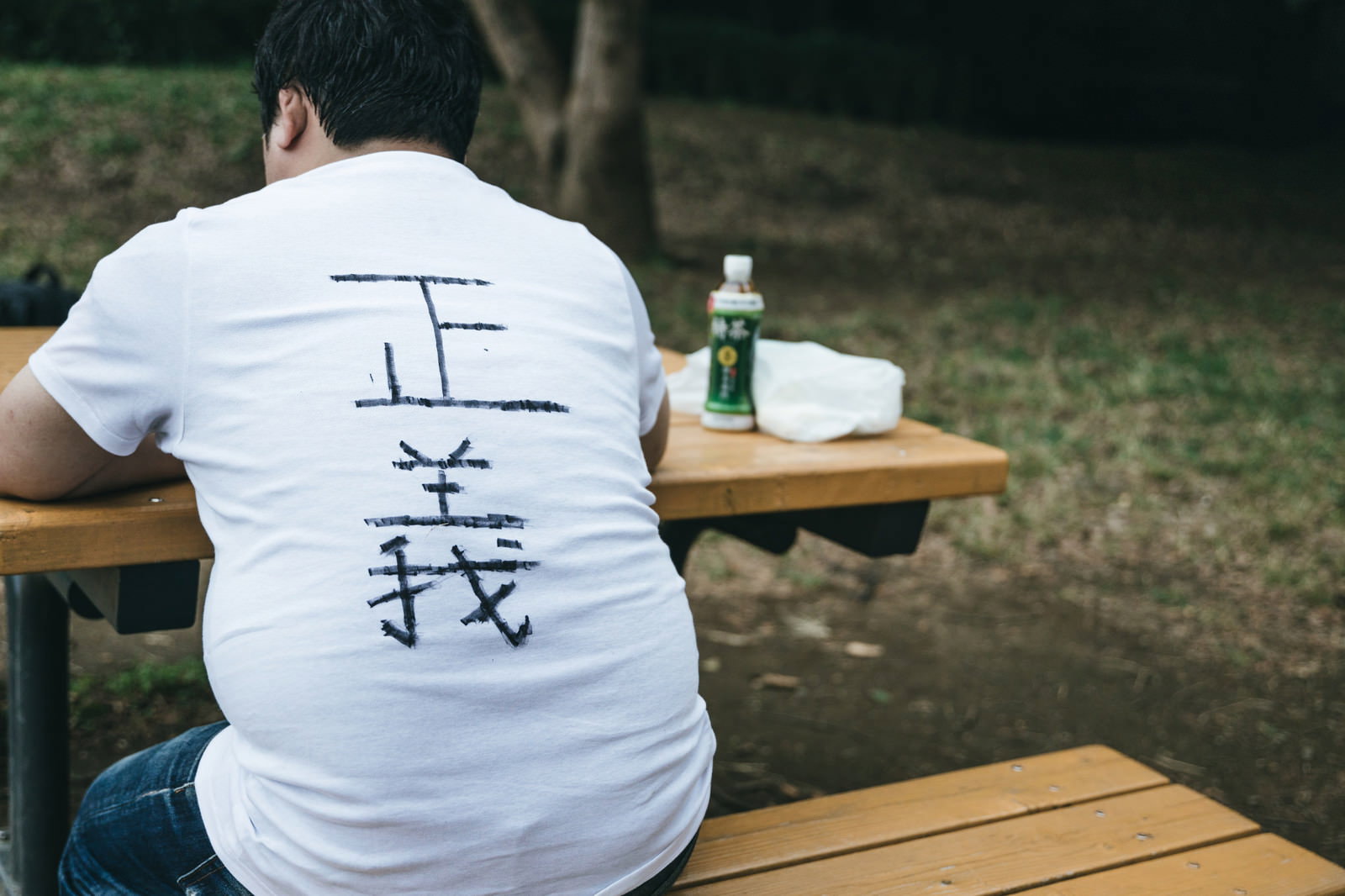 正義と書かれた白いTシャツを着て木製テーブルに座る人の後ろ姿［モデル：段田隼人］
