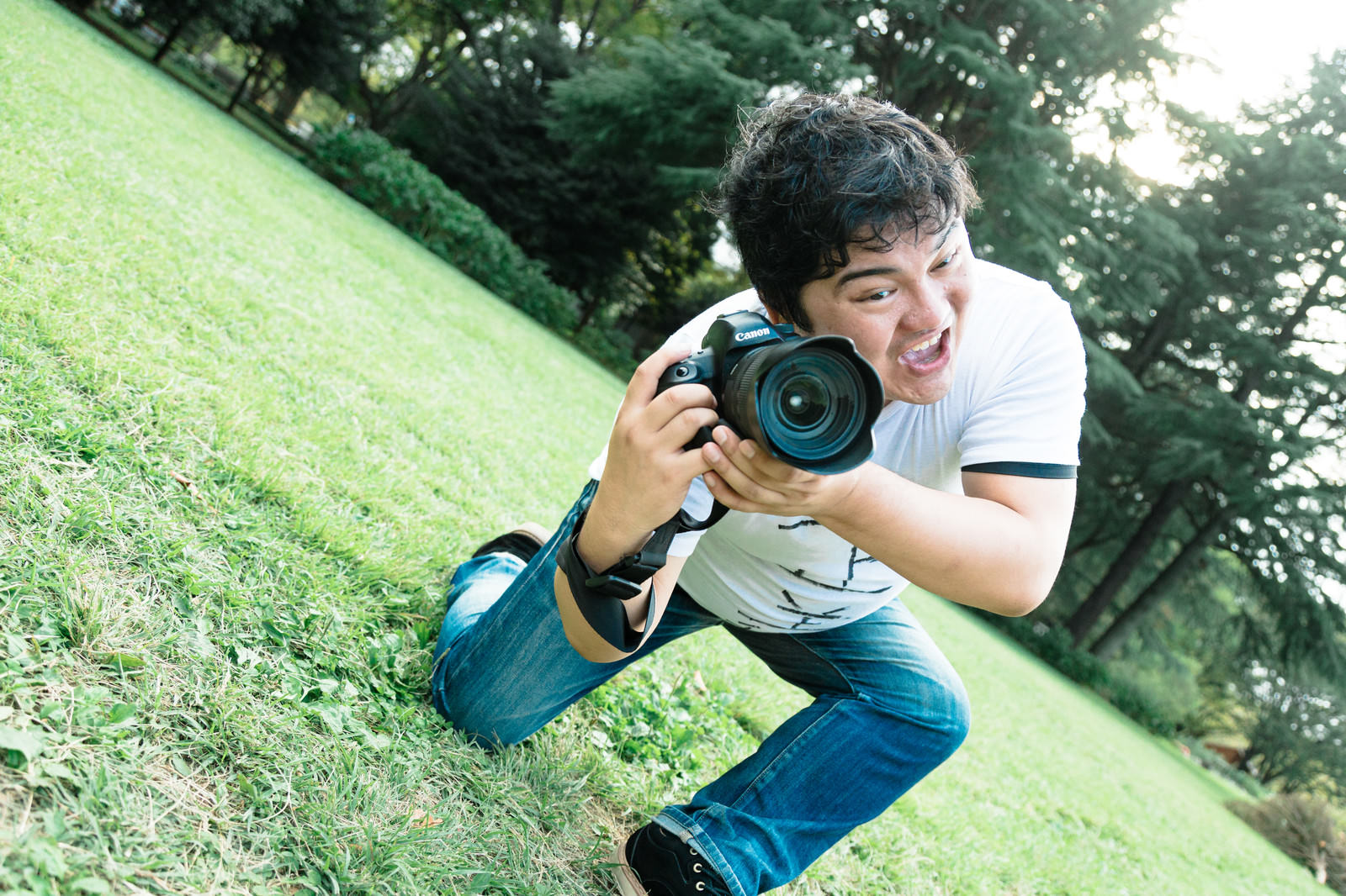 白いTシャツを着た男性がカメラを構えて芝生で撮影している様子［モデル：段田隼人］