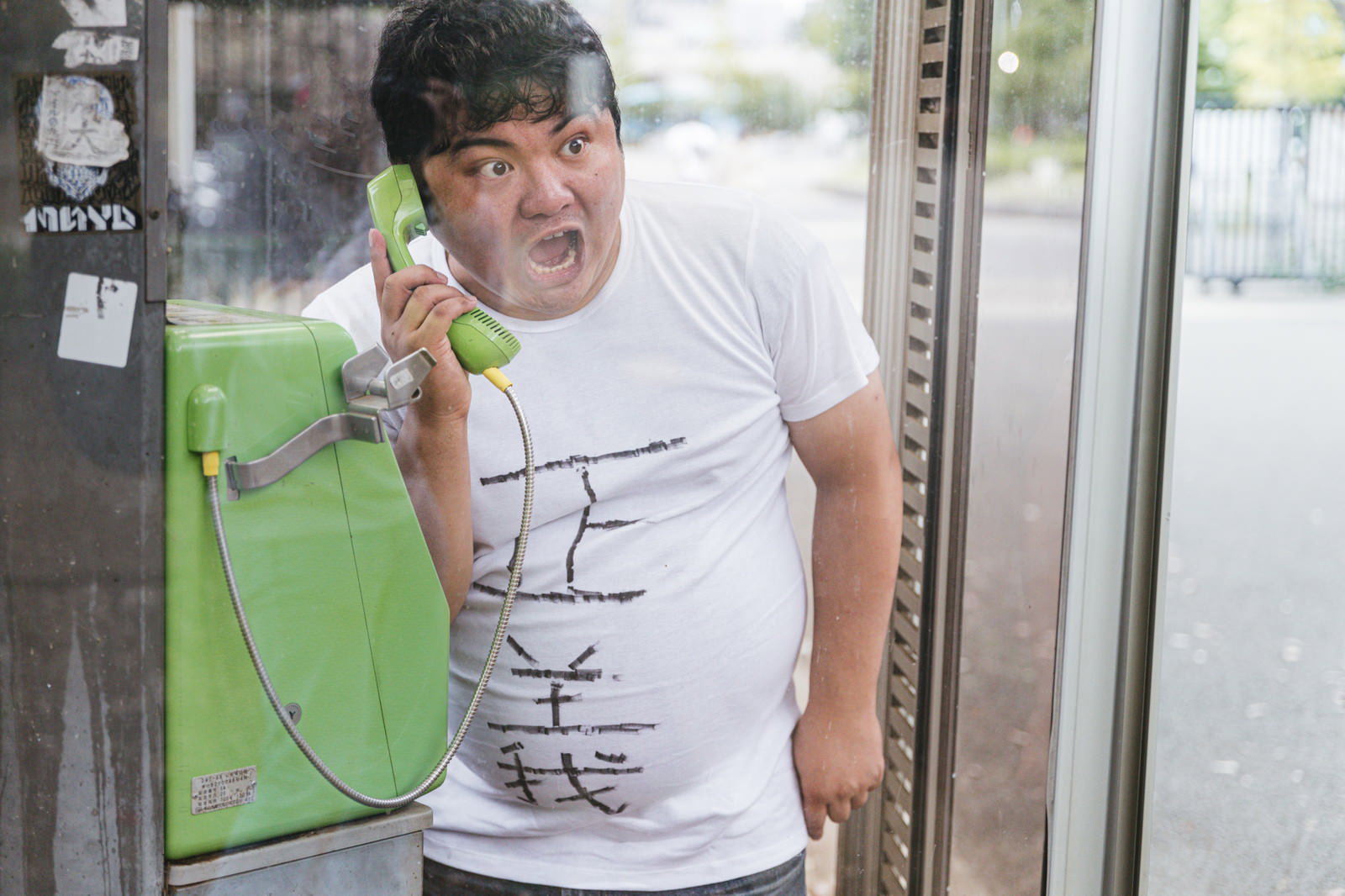 公衆電話ボックスで怒った表情で通報の電話をかける男性［モデル：段田隼人］