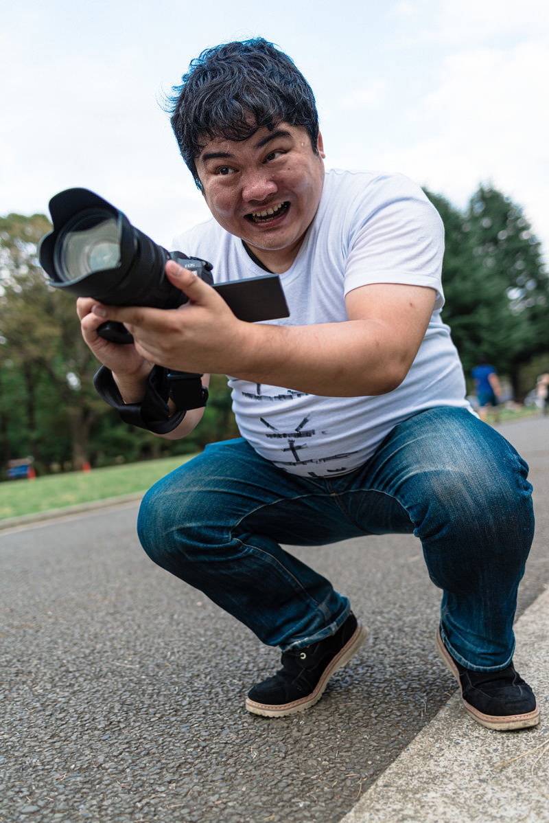 道路でしゃがんでローアングルからカメラを構える白いTシャツの男性［モデル：段田隼人］
