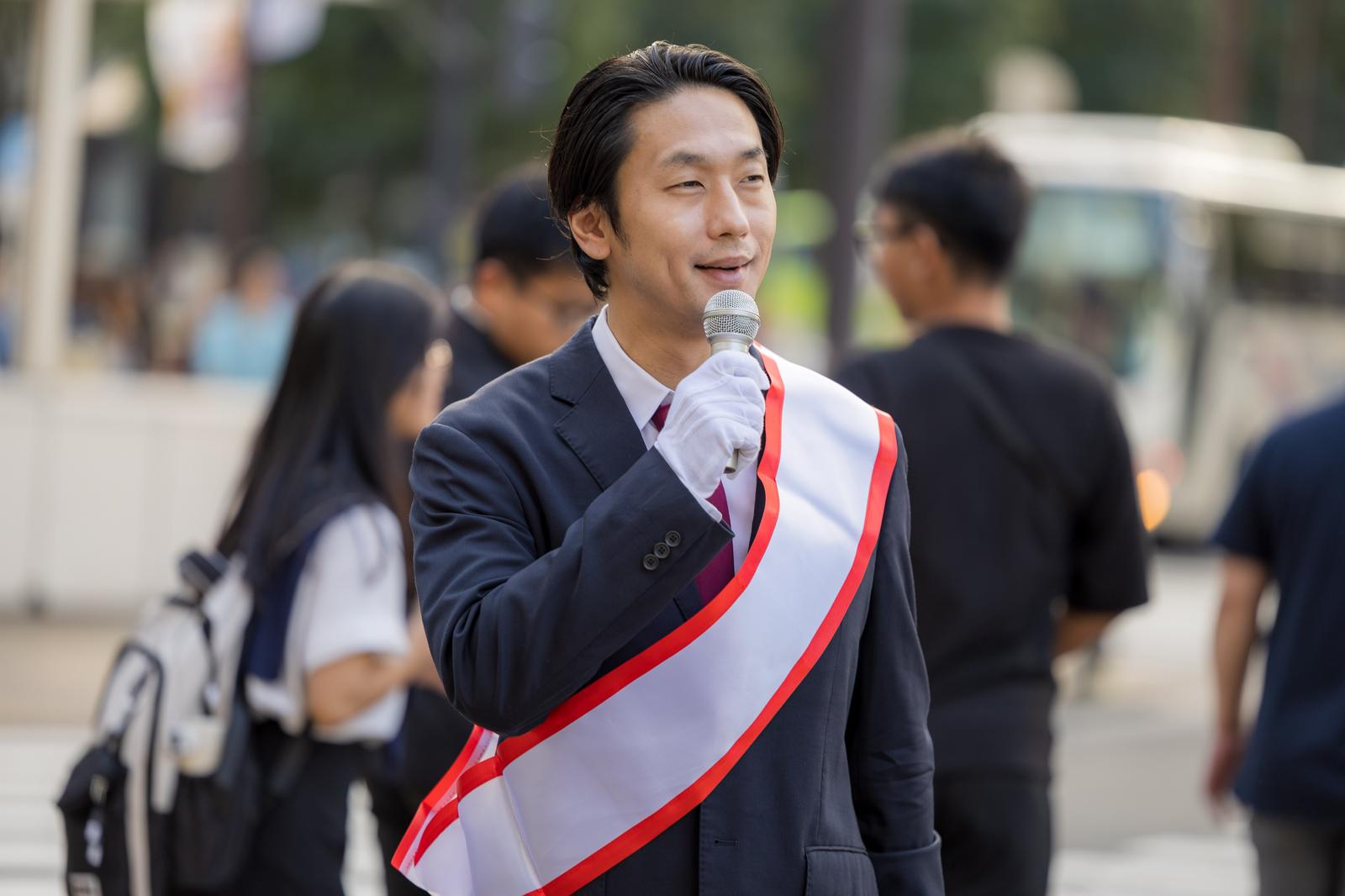 街頭でマイクを持ってタスキをかけて演説する男性政治家［モデル：大川竜弥］