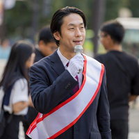 街頭でタスキをかけて演説する男性政治家のマイク演説の写真