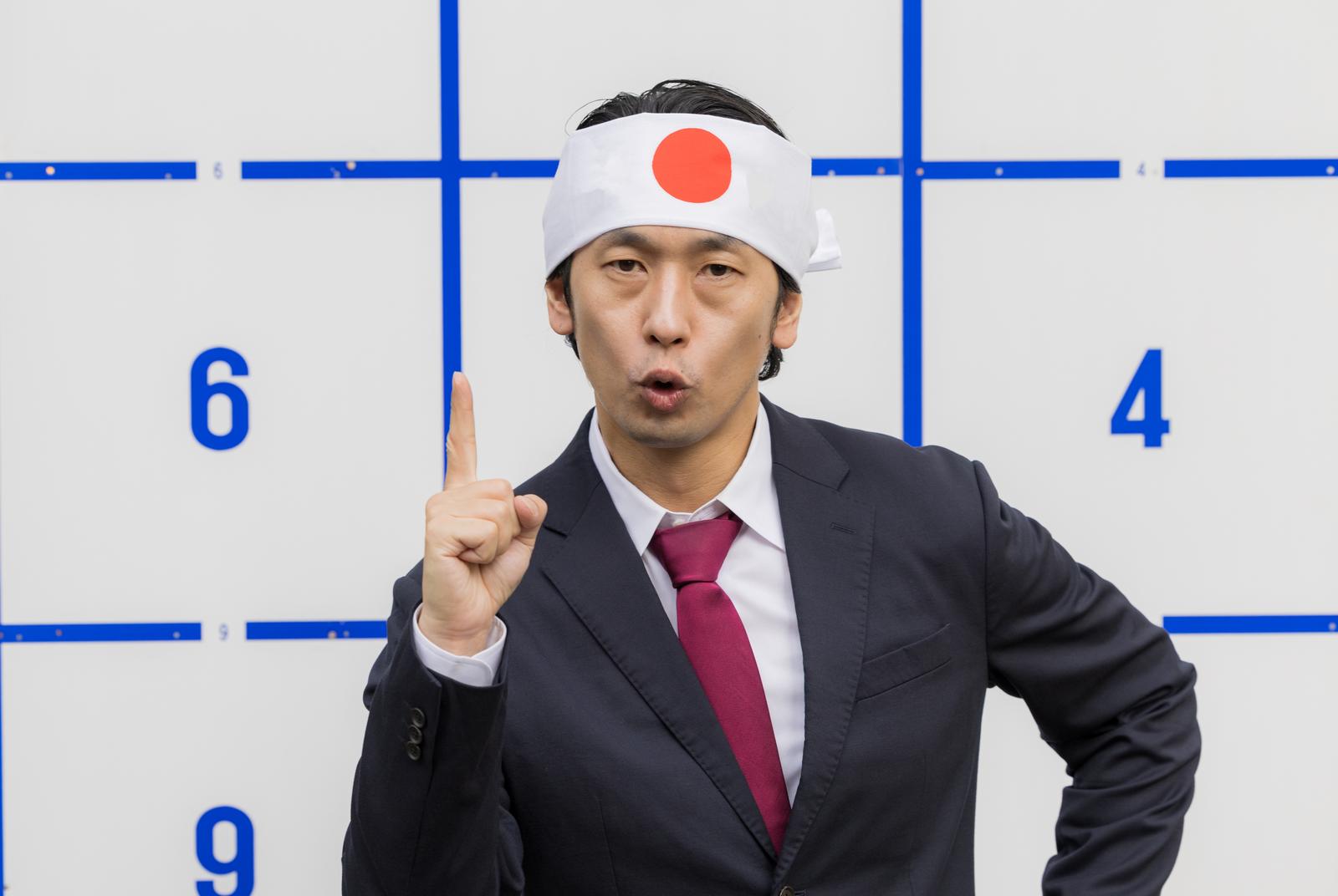 日の丸の鉢巻を巻き、ナンバーワンを掲げるスーツ姿の男性［モデル：大川竜弥］