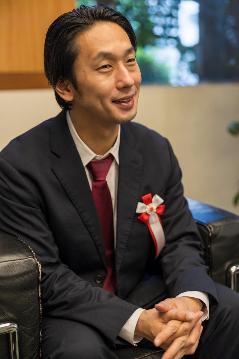 赤白リボンバッジを付けた紺スーツの男性政治家が、笑顔で右側に向いて談話している［モデル：大川竜弥］
