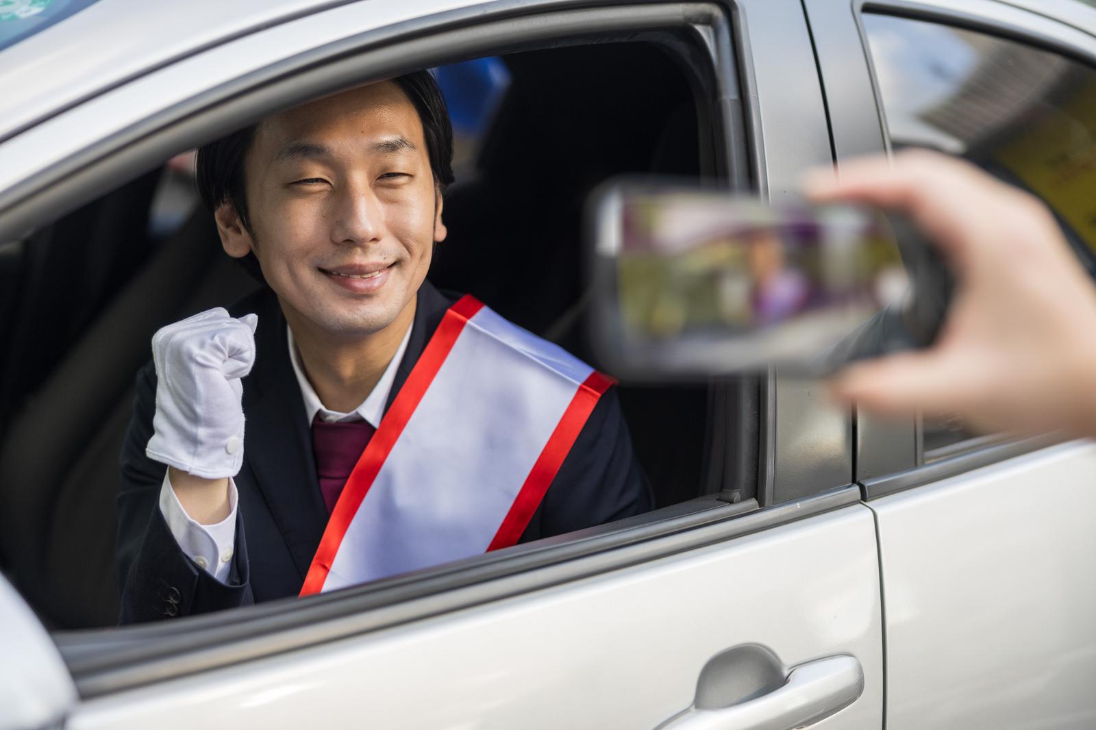 車窓から笑顔で演説する、たすきを掛けたスーツ姿の男性政治家［モデル：大川竜弥］