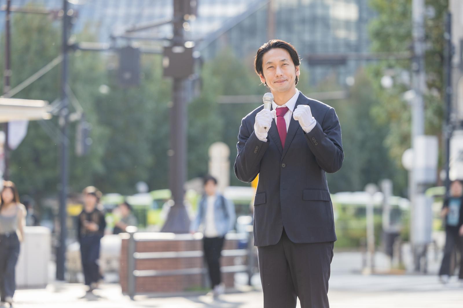 駅前でマイクを握り話す、スーツ姿の政治家風の男性［モデル：大川竜弥］