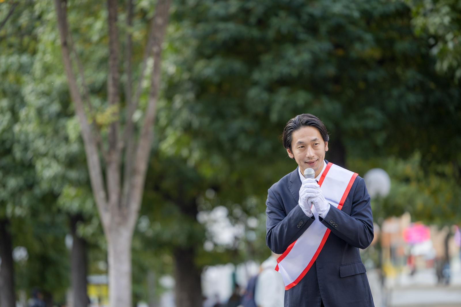 マイクを握り街頭演説する赤白のたすきをかけた男性政治家［モデル：大川竜弥］