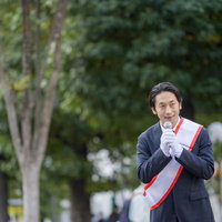 緑豊かな公園でマイクを握り街頭演説する若い男性政治家の写真