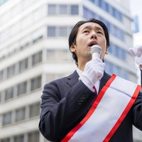 たすきを着けて街頭演説するビジネススーツ姿の40代男性の写真