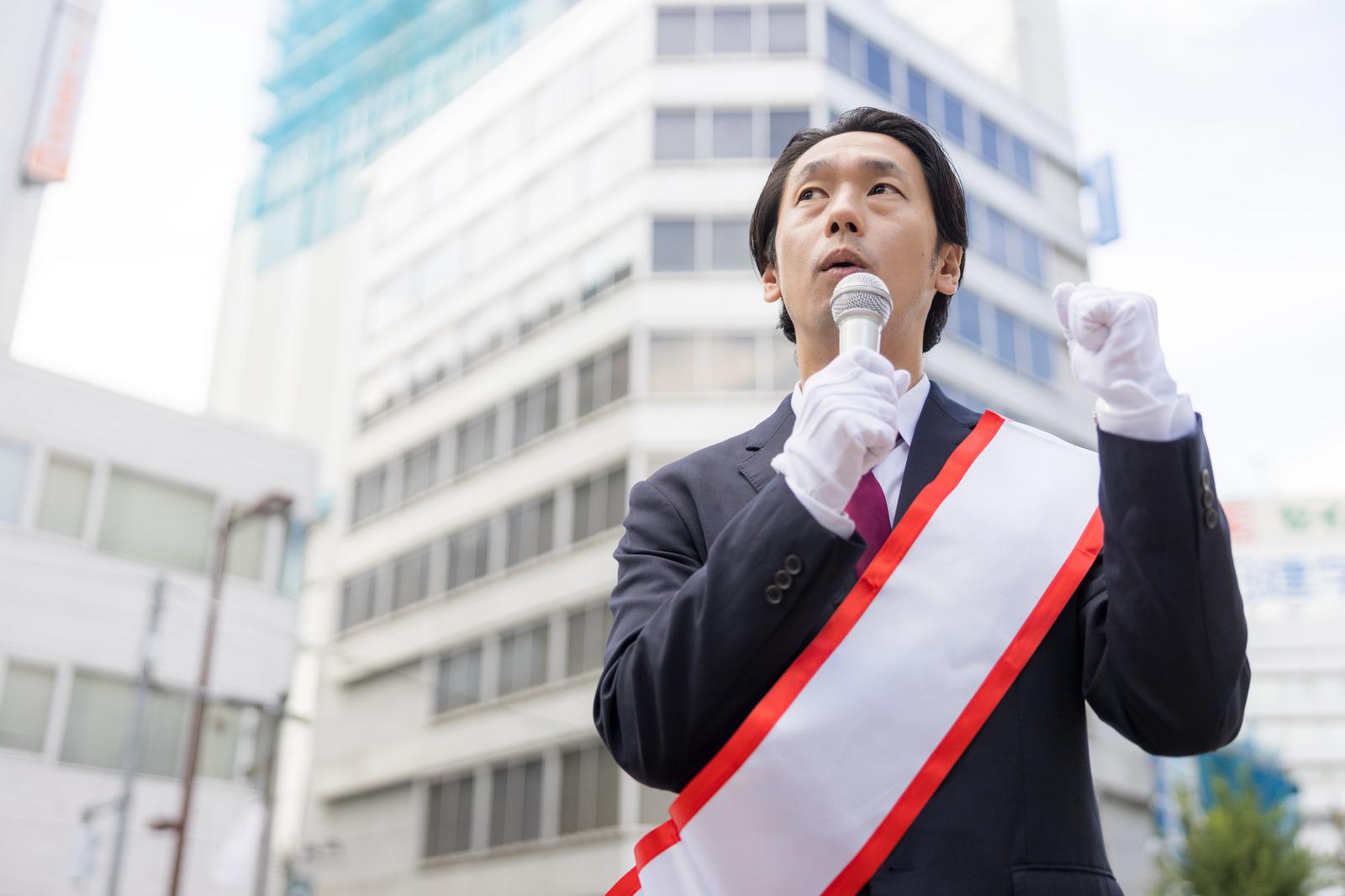 たすきを掛けた男性が上を見ながらマイクで街頭演説をしている［モデル：大川竜弥］