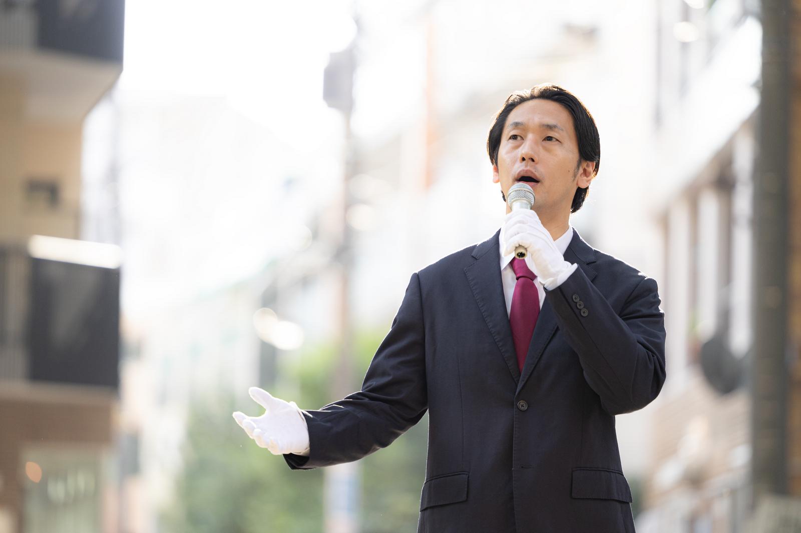スーツ姿でマイクを持ち、住宅街で街頭演説する政治家風の男性［モデル：大川竜弥］
