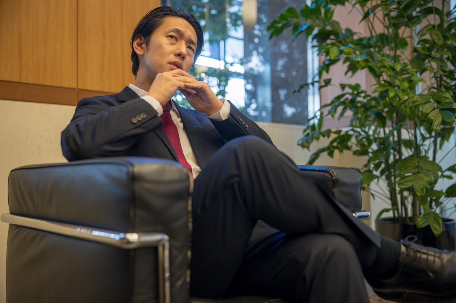 ソファーで顎に手を当てて思案する紺スーツの男性［モデル：大川竜弥］