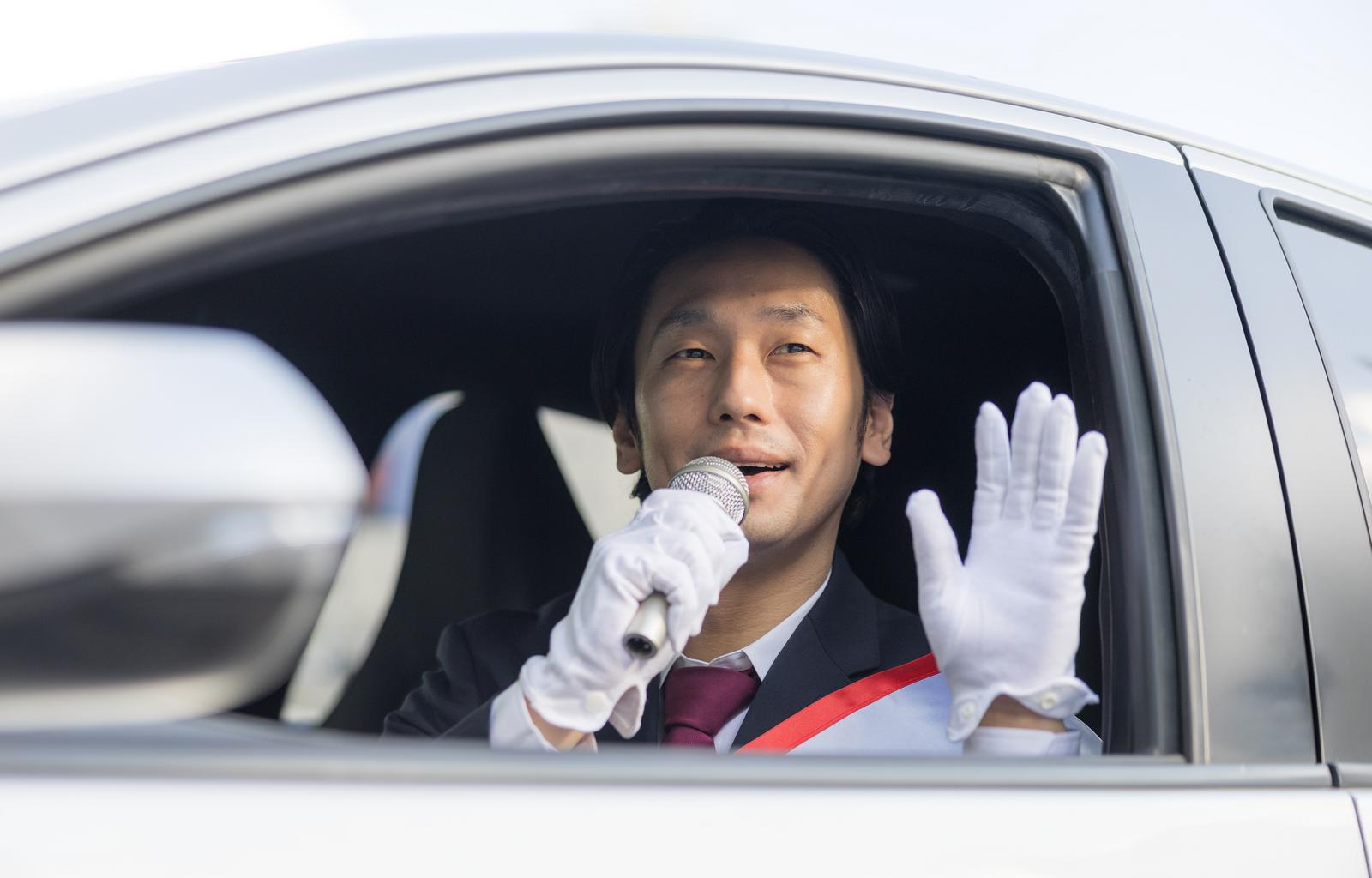 車の窓からマイクを持って右手を上げて手を振る選挙人の男性［モデル：大川竜弥］