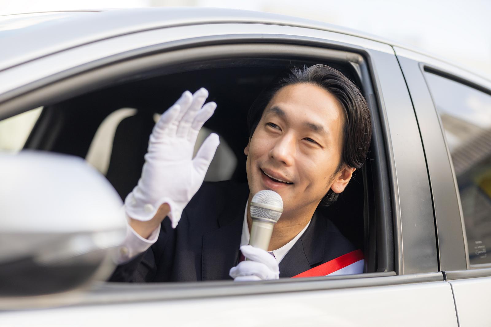 タスキを掛けたスーツ姿の男性がマイクを持ち、車窓から手を振る［モデル：大川竜弥］