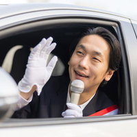 車の窓から顔を出してマイクで演説する政治家風の男性の写真