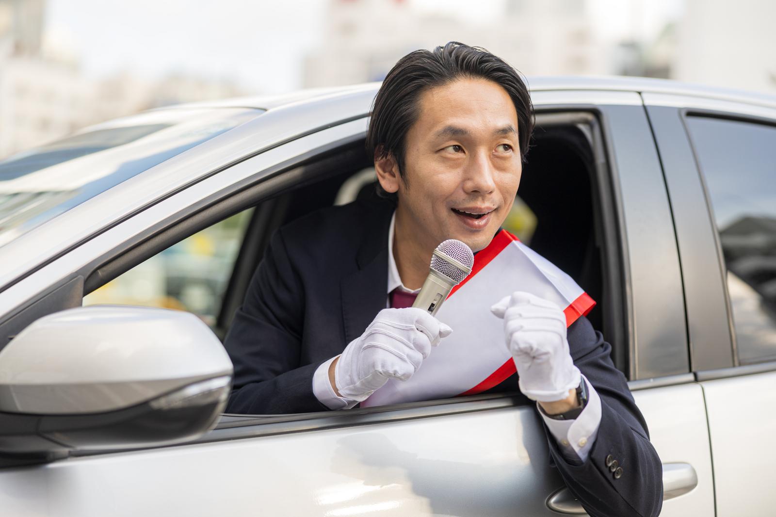 車の窓から身を乗り出してマイクで演説する男性［モデル：大川竜弥］
