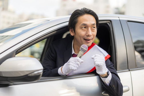 車窓からマイクで身を乗り出してアピールする政治家風の男性