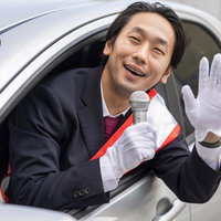 停車中の白い車から身を乗り出してマイクで演説する男性政治家の写真