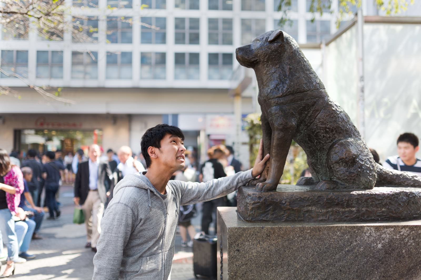 渋谷の人混みの中、グレーのパーカーを着た男性がハチ公像に手を当てながら上を向いて笑っている［モデル：大川竜弥］