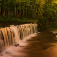 森林に囲まれた滝の流れと白い水しぶき、夕暮れの光に照らされた水面の写真
