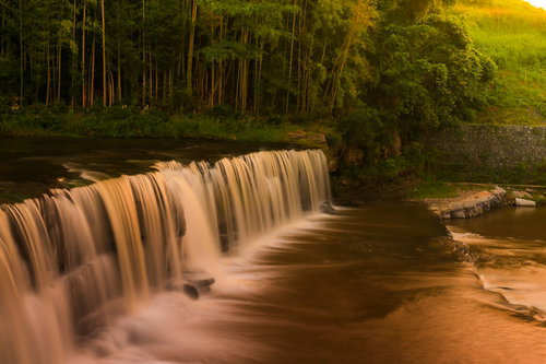 森林に囲まれた滝の流れと白い水しぶき、夕暮れの光に照らされた水面