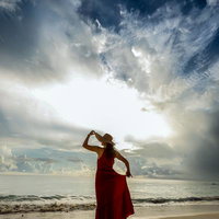 海辺でハットを手に持つ赤いドレスの女性が空を見上げるポートレートの写真