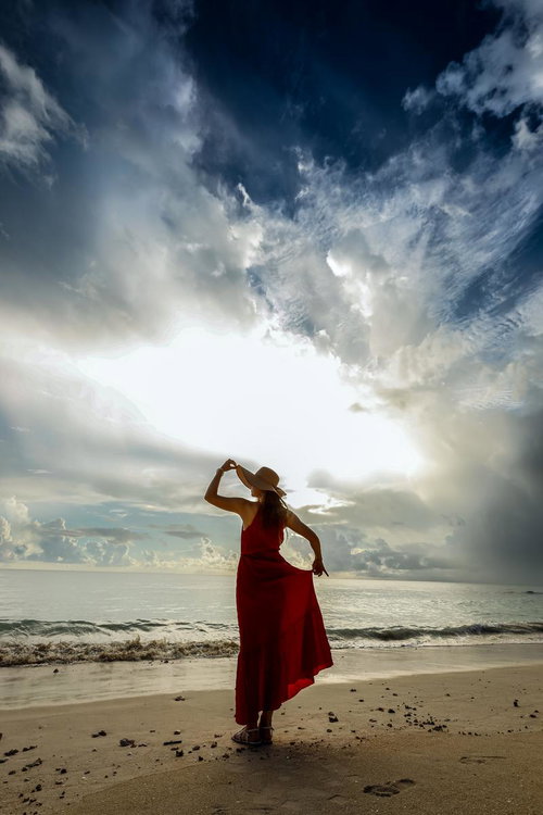 海辺でハットを手に持つ赤いドレスの女性が空を見上げるポートレート