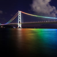 虹色にライトアップした夜の明石海峡大橋と海面の光景の写真