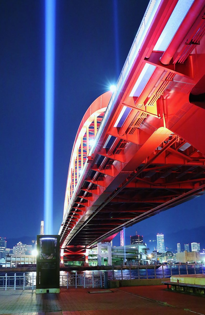 赤くライトアップされた神戸大橋を見上げたアングルで撮影し、青いレーザービームが夜空を照らしている