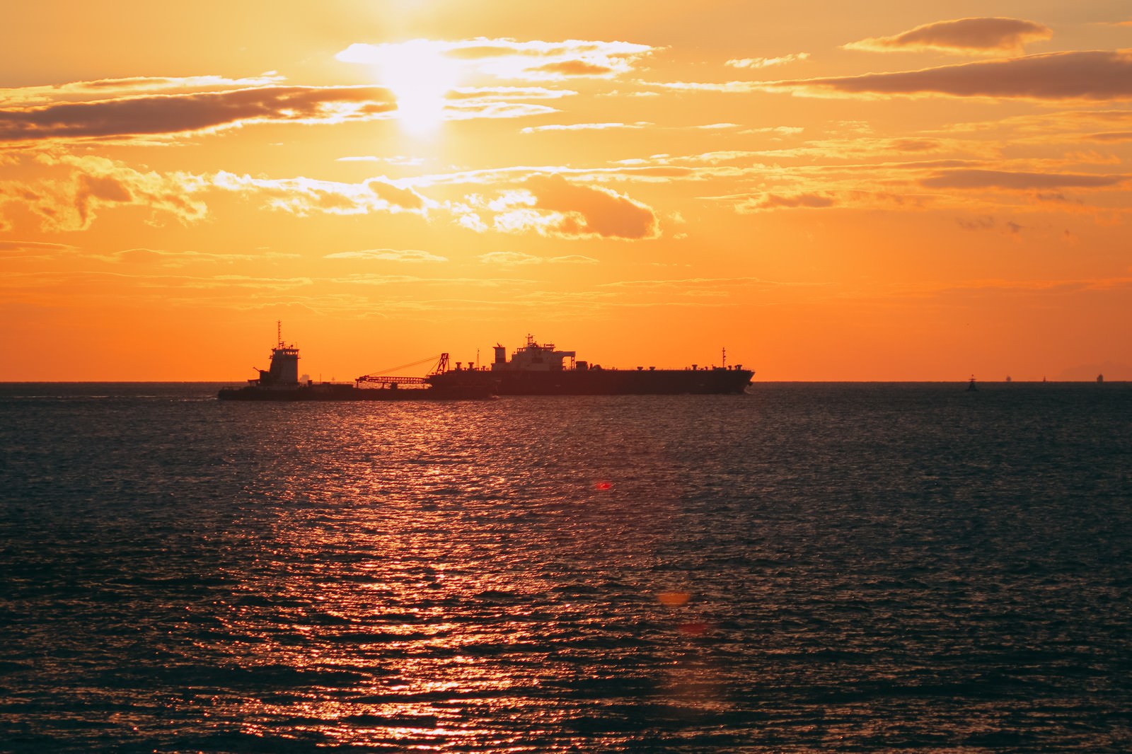 オレンジ色の夕焼け空の下、海面のさざ波が輝き遠くに船のシルエットが見える海の風景