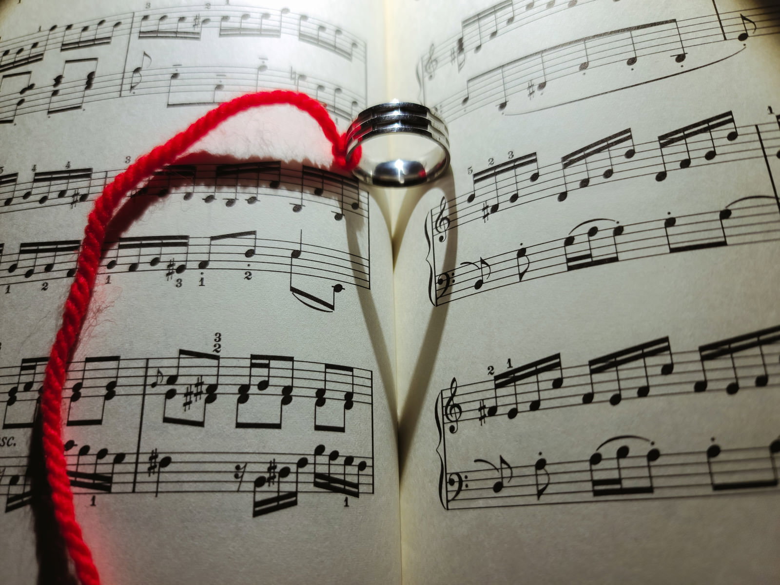 楽譜の上に置かれた指輪から赤い糸が伸び、ハート型の影が映る