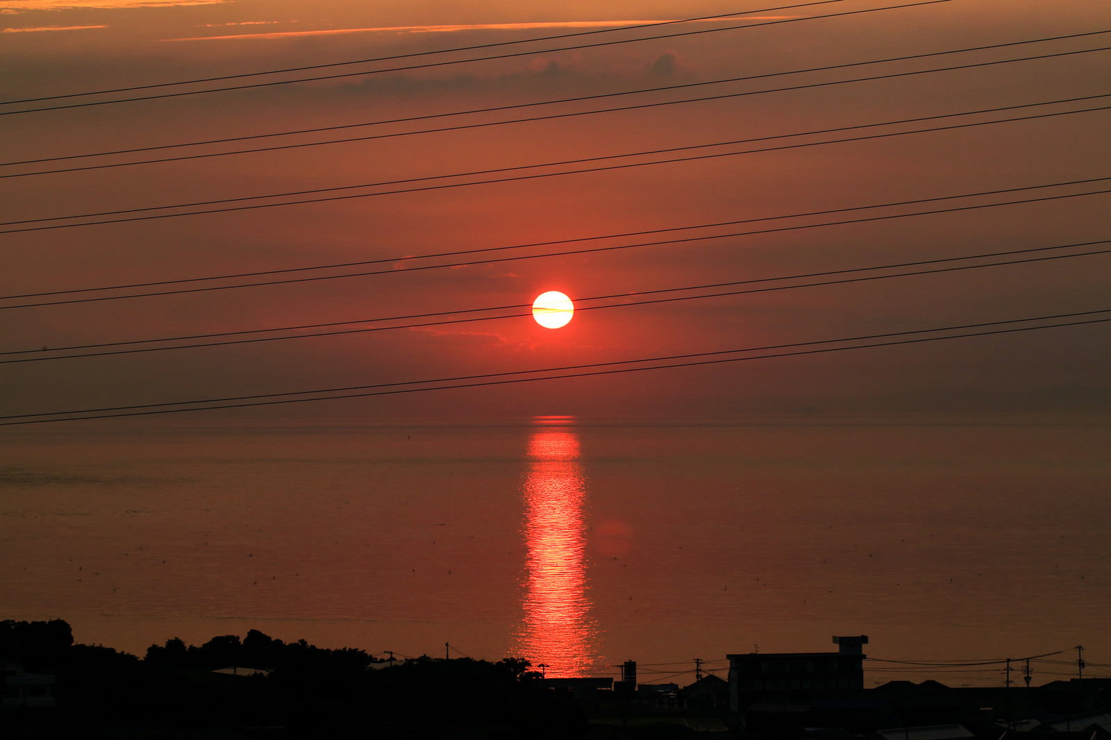 オレンジ色の夕焼け空の下、水平線に沈む赤い太陽とその光が海面に映るレイライン