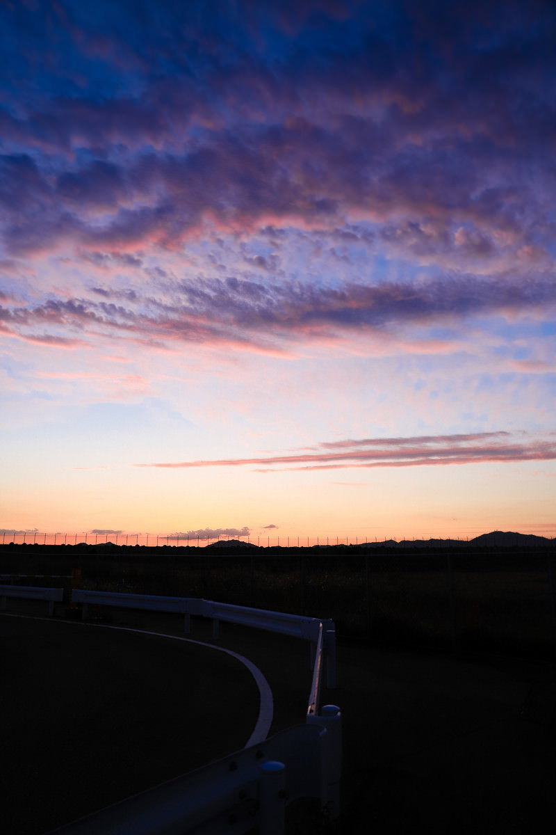 紫とピンクに染まった夕焼け空の下、暗い大地に続く一本の田舎道