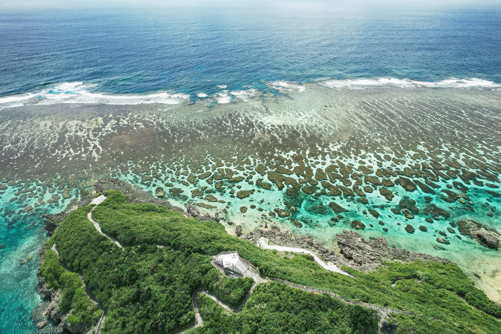 宮古島の岩礁と透明な海を空撮した写真。手前に緑に覆われた岩場と砂浜、奥に波が寄せる海岸線が広がる