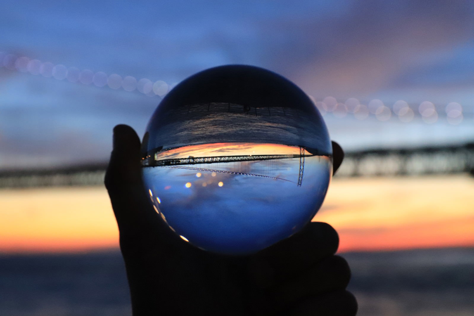 手に持ったガラス玉に夕焼け空と橋が反転して映り込んでいる写真