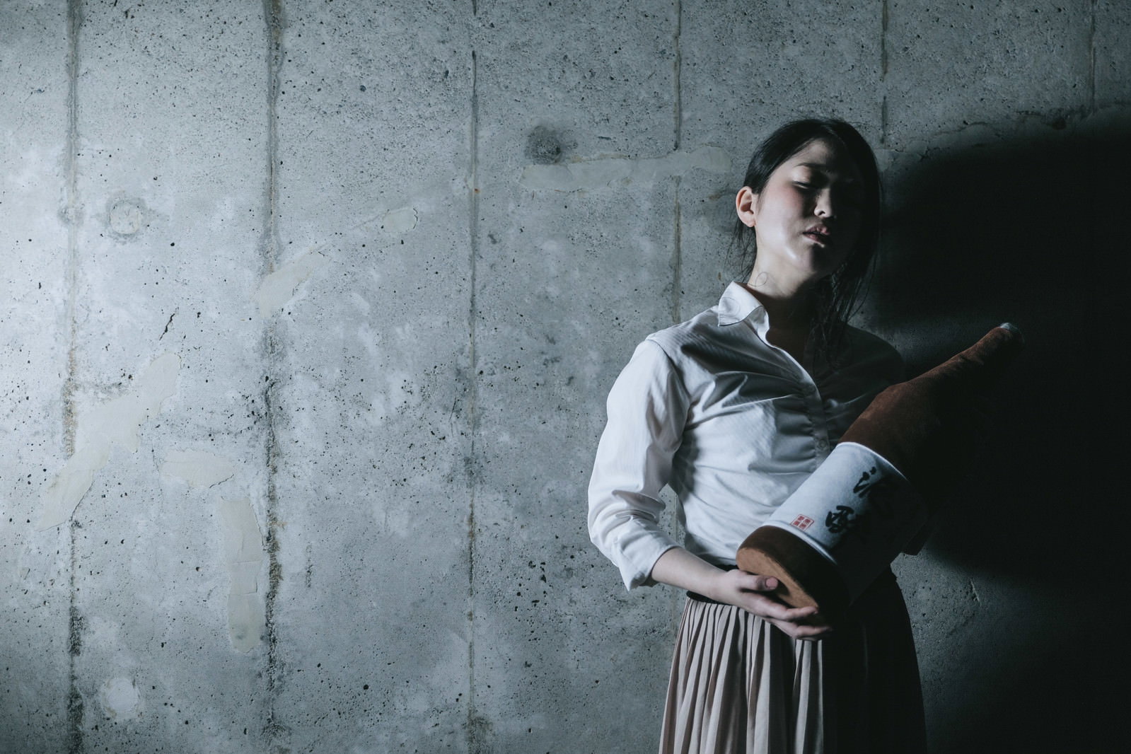 コンクリートの壁際に座り込み、目を閉じてうつむいているシャツ姿の女性［モデル：シマヅ］