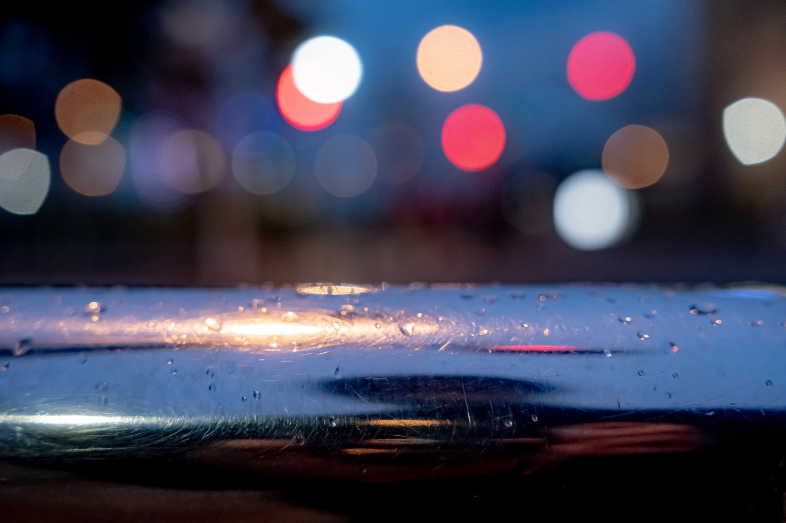 手すりに付いた雨粒と背景にぼけた赤白のライトが玉ボケ状に浮かぶ夜景