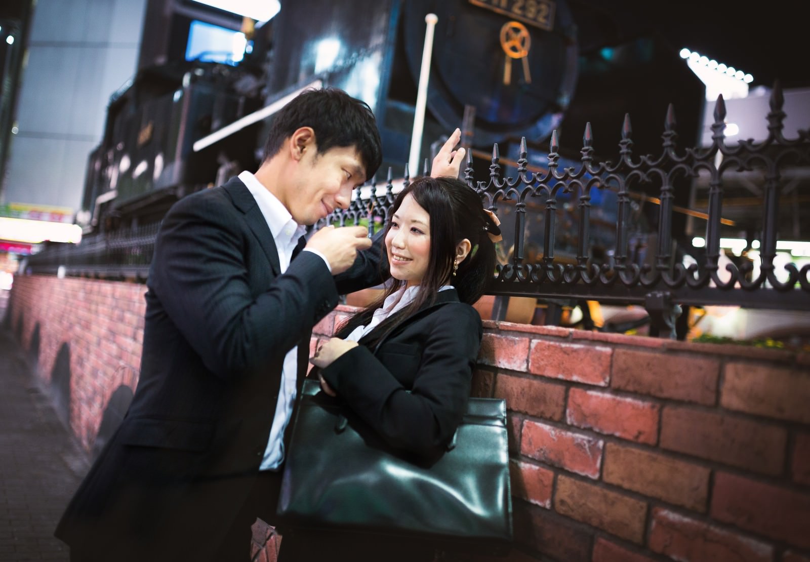 夜間の駅前でスーツ姿の男性が女性に身を寄せながら話しかけ、背景に黒い柵と夜間の街灯が見える［モデル：大川竜弥 シマヅ］
