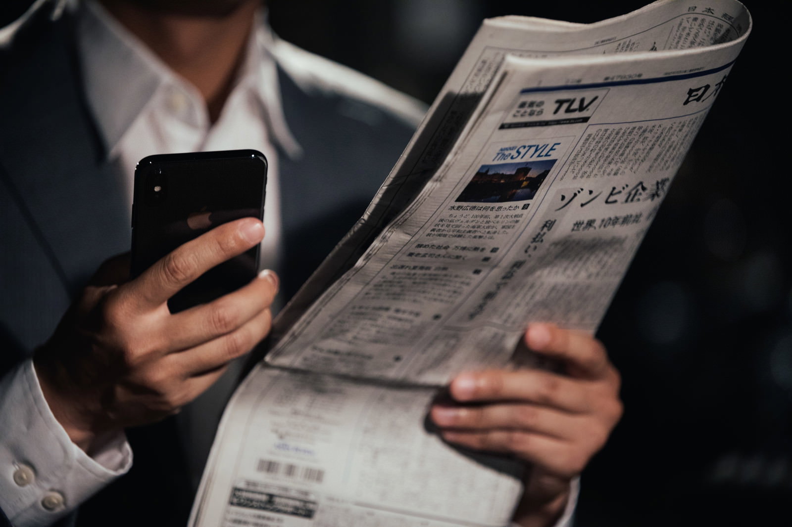 新聞を片手にスマートフォンを操作するスーツ姿のビジネスマン［モデル：大川竜弥］