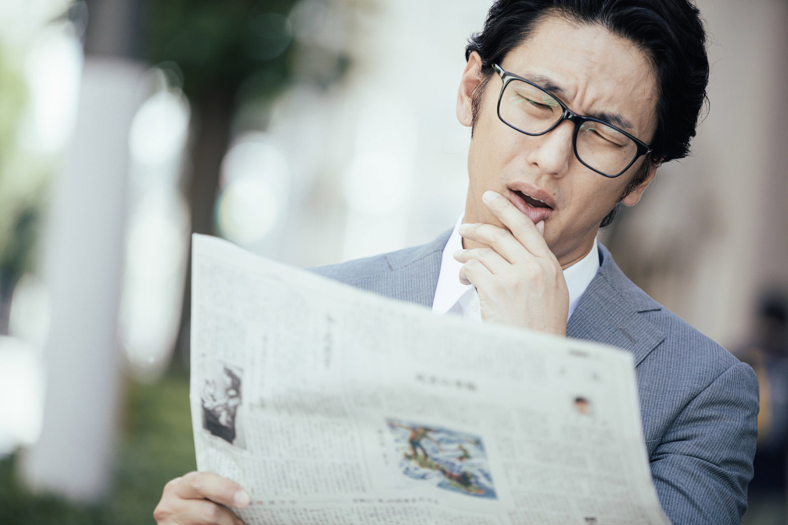 新聞を読んで手で口を覆って驚いている眼鏡をかけた男性［モデル：大川竜弥］