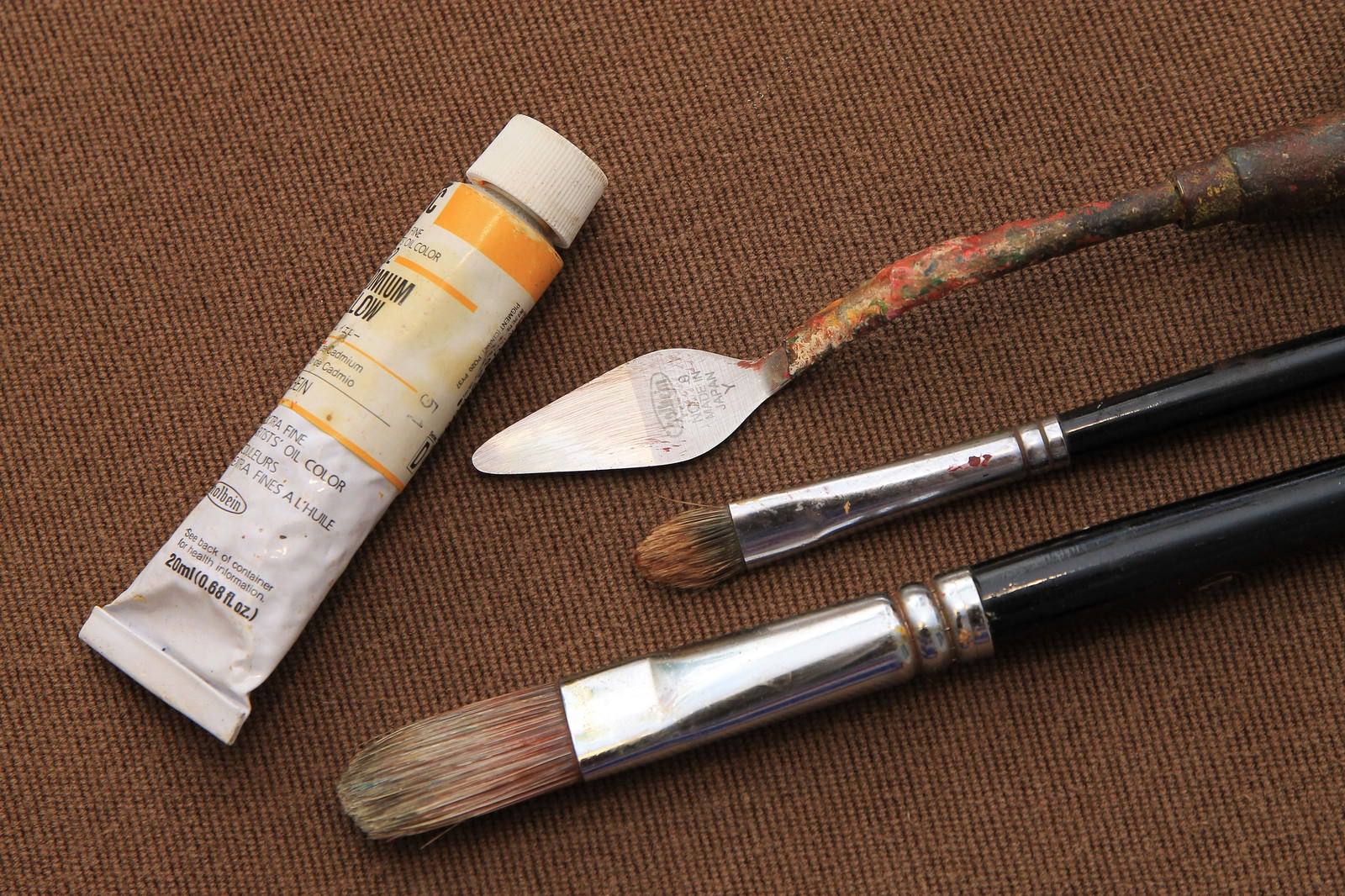 油絵の絵の具、パレット、筆が茶色い布の上に並べられている